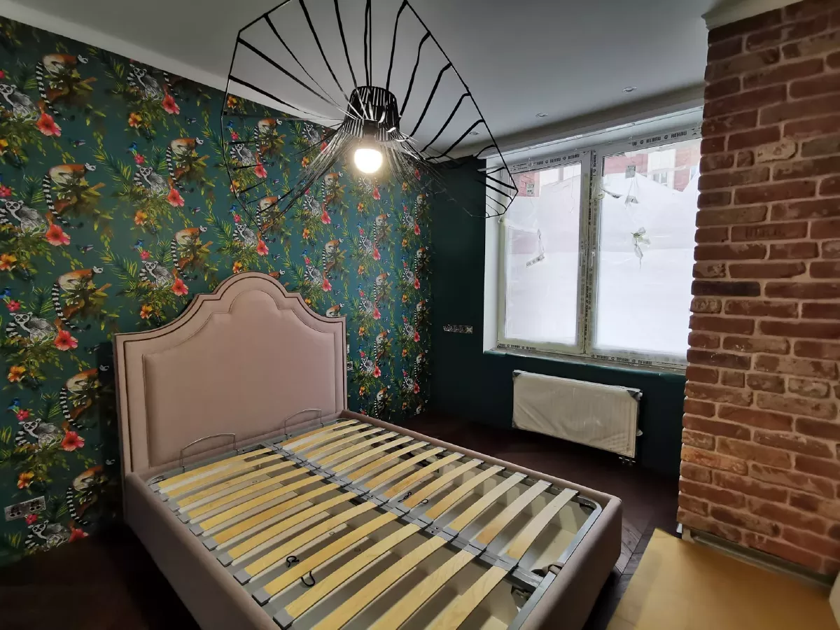 Дизайнерский ремонт спальни: собран каркас двуспальной кровати, стена из декоративного кирпича, обои с экзотическим принтом, паркетная доска французская елка, двухслойный гипсокартонный потолок с гипсовыми молдингами и плинтусами