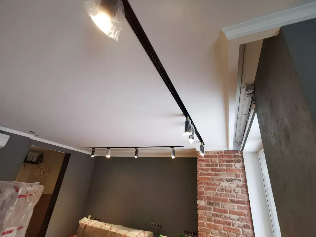 Дизайнерская отделка гостиной в новостройке: трековое освещение на потолке из гипсокартона, ниши под скрытые карнизы, гипсовые молдинги по периметру, декоративная штукатурка темно-серого цвета, колонна из декоративного кирпича, кондиционер