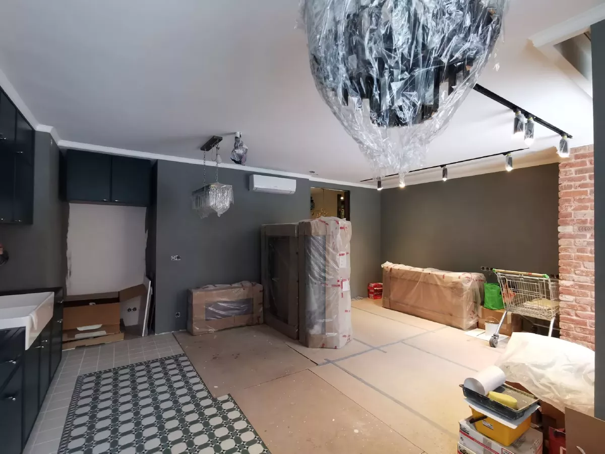 Ремонт кухни-гостиной в новостройке: декоративная штукатурка темно-серого цвета, трековое освещение, ниши под скрытые карнизы, акцентная стена из декоративного кирпича, геометрическая плитка в зоне кухни, пол защищен ДВП, дизайнерский интерьер